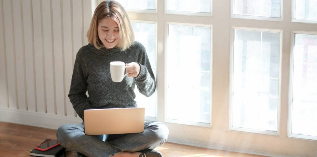 A woman sitting by a tall window with a laptop.